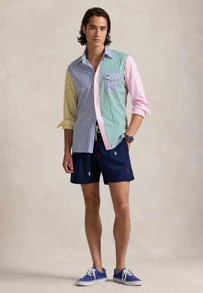 Πουκάμισο με κουμπιά και οριζόντιες ρίγες σε πολλά χρώματα με αποχρώσεις του πράσινου, ροζ και μπλε· συνδυασμένο με ναυτικά μπλε μαγιό σορτς με μικρά λογότυπα.