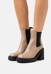 Bottines en cuir beige avec des panneaux élastiques noirs sur les côtés et des talons larges à crampons noirs. Texture lisse, bout rond et détail de tirette.