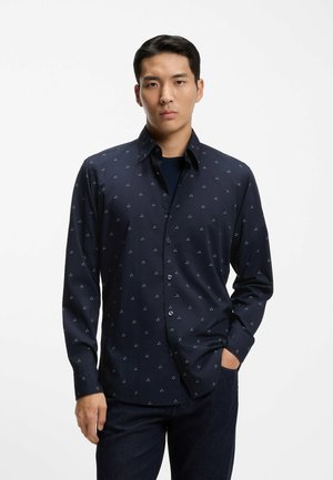 Camicia blu navy con piccoli motivi a triangolo bianco, maniche lunghe, colletto con bottoni, tessuto liscio e leggermente lucido. Indossata sopra un denim scuro.