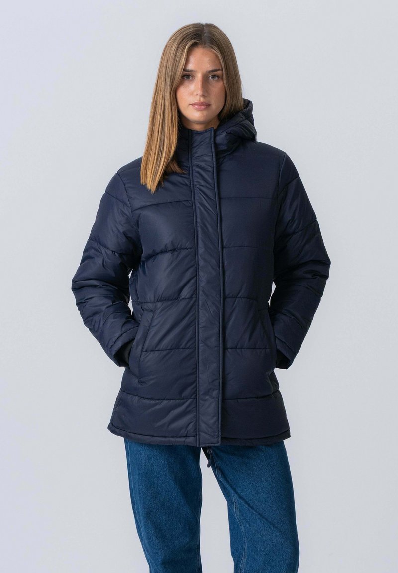 Ung kvinde iført en marineblå polstret hættejakke med hænderne i lommerne og blå jeans, stående foran en ensfarvet lys grå baggrund.