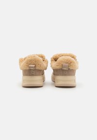Sneakers beige con fodera soffice di colore crema, materiale scamosciato e suola modellata. Etichetta con logo sul tallone. Design minimalista.