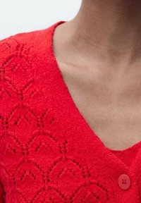Rød strikket sweater med V-udskæring, indviklet diamantmønster og en afrundet knapdetalje på venstre side nær halsudskæringen.