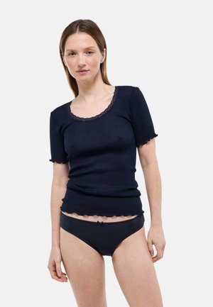 Femme portant un haut moulant côtelé bleu marine à manches courtes avec bordure en dentelle et une lingerie assortie bleu marine, debout devant un fond clair uni.
