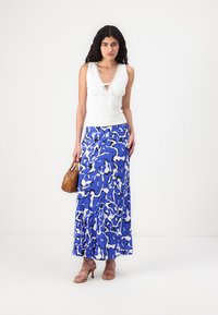 Hvit ermeløs topp med blondekant, kombinert med et langt, flytende blått skjørt med hvite og mørkeblå abstrakte mønstre. Brun håndveske og høye hæler.