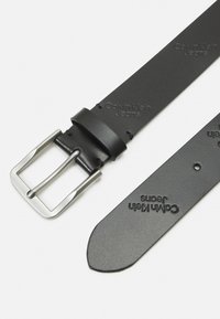 Cintura in pelle nera con una fibbia in metallo argentato. Presenta il logo "Calvin Klein Jeans" in rilievo sulla cintura. Texture liscia, design classico.