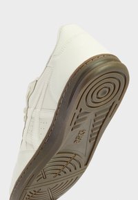 Bílá kožená teniska s kulatou špičkou, texturovanými detaily a gumovou podrážkou se spirálovým vzorem a jemným logem na patě.