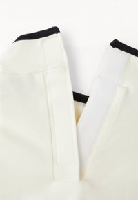 Cream v-neck top with black trim, made of ribbed fabric. Features a placket with hook-and-loop closure for adjustable fit and design.