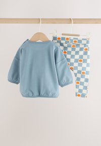 Blue long-sleeve baby shirt and checkered blue and white pants with orange pumpkin print hanging on wooden hangers on a wooden rod.