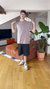 Mlad čovjek u bordo kapi drži s bočnih strana svijetloplavog majicu, stojeći u dnevnoj sobi s kaučem, biljkom i televizorom u pozadini.