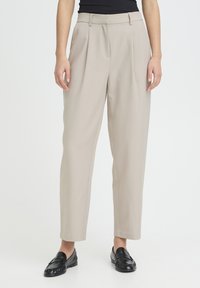 Pantalon beige sur mesure à taille haute, avec des plis et des jambes droites. Confectionné dans un tissu lisse, associé à des mocassins noirs.