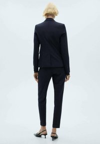 Costume marine avec un blazer ajusté à une fente arrière, un pantalon slim et des talons noirs pointus, présenté de dos.