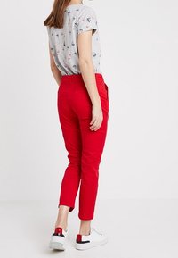 Rode slim-fit broek met een gladde textuur, gecombineerd met een lichtgrijze shirt met een subtiel nautisch patroon. Witte sneakers met accenten.