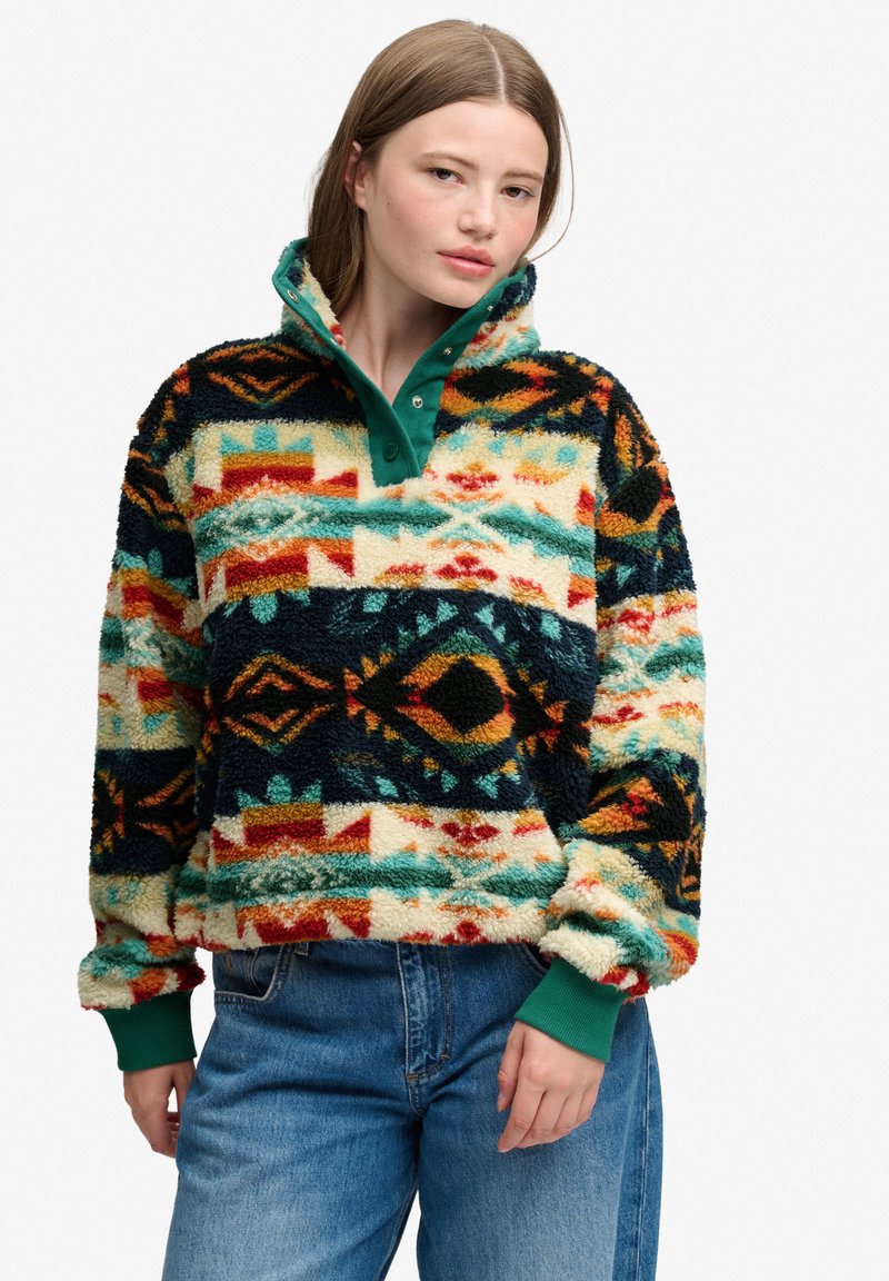 Femme portant un pull polaire à motifs colorés avec col et poignets verts, assorti à un jean bleu, debout devant un fond uni.