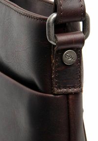 Borsa a mano in pelle marrone che mostra un primo piano della tracolla con una fibbia in metallo argentato e un dettaglio a bottone. Superficie texturizzata con cuciture.