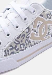 Άσπρο αθλητικό παπούτσι με γκρι και μπεζ λεοπάρ σχέδιο, μπεζ λογότυπο DC, άσπρα κορδόνια και μπεζ ρίγα στη λευκή καουτσούκ σόλα.