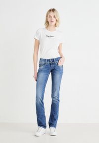 T-shirt blanc avec logo noir, associé à un jean en denim bleu légèrement décoloré et à une braguette à deux boutons, complété par des baskets blanches.