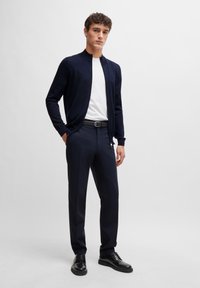 Navy cardigan over a white t-shirt, paired with navy trousers and black lace-up shoes. The cardigan features a collarless design.