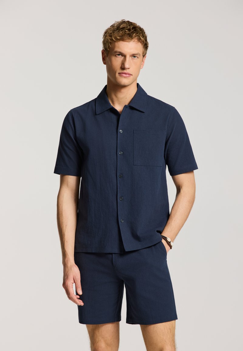 Mann trägt marineblaue kurzärmelige Hemdbluse und passende Shorts, steht mit einer Hand in der Tasche vor einfachem Hintergrund.