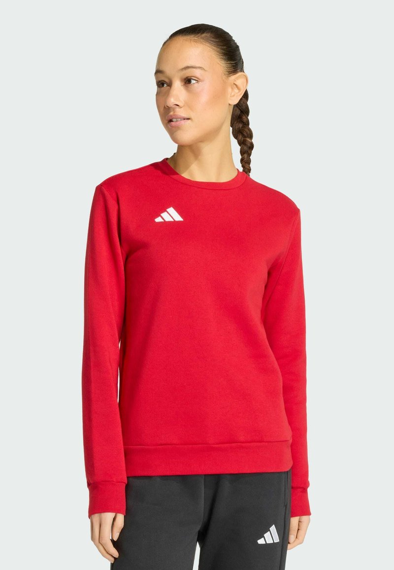 Femme aux cheveux tressés portant un sweat-shirt rouge et un pantalon noir, tous deux avec un logo Adidas blanc, debout devant un fond uni.