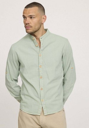 Chemise vert clair à manches longues avec un col mandarin, des boutons couleur beige et des manches retroussées. Tissu doux avec une texture lisse et un léger éclat.