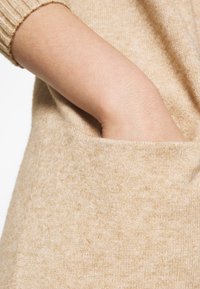 Beige stickad plagg med en stor framficka, mjuk textur, ribbad ärmavslut och en avslappnad passform.