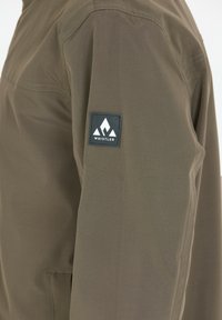 Oliivinvihreä takki, jossa on sileä pinta, rinnassa logo-patch ja hienovaraisia ompeleita. Yksinkertainen, nykyaikainen muotoilu.