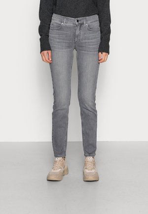 Person iført grå slim-fit jeans, beige sneakers og en mørkegrå langærmet sweater, stående mod en ensfarvet baggrund.