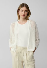 Witte gebreide cardigan met open gaasdesign, gecombineerd met een witte tanktop en lichtbeige broek. Heeft een relaxte pasvorm en ribgebreide manchetten.