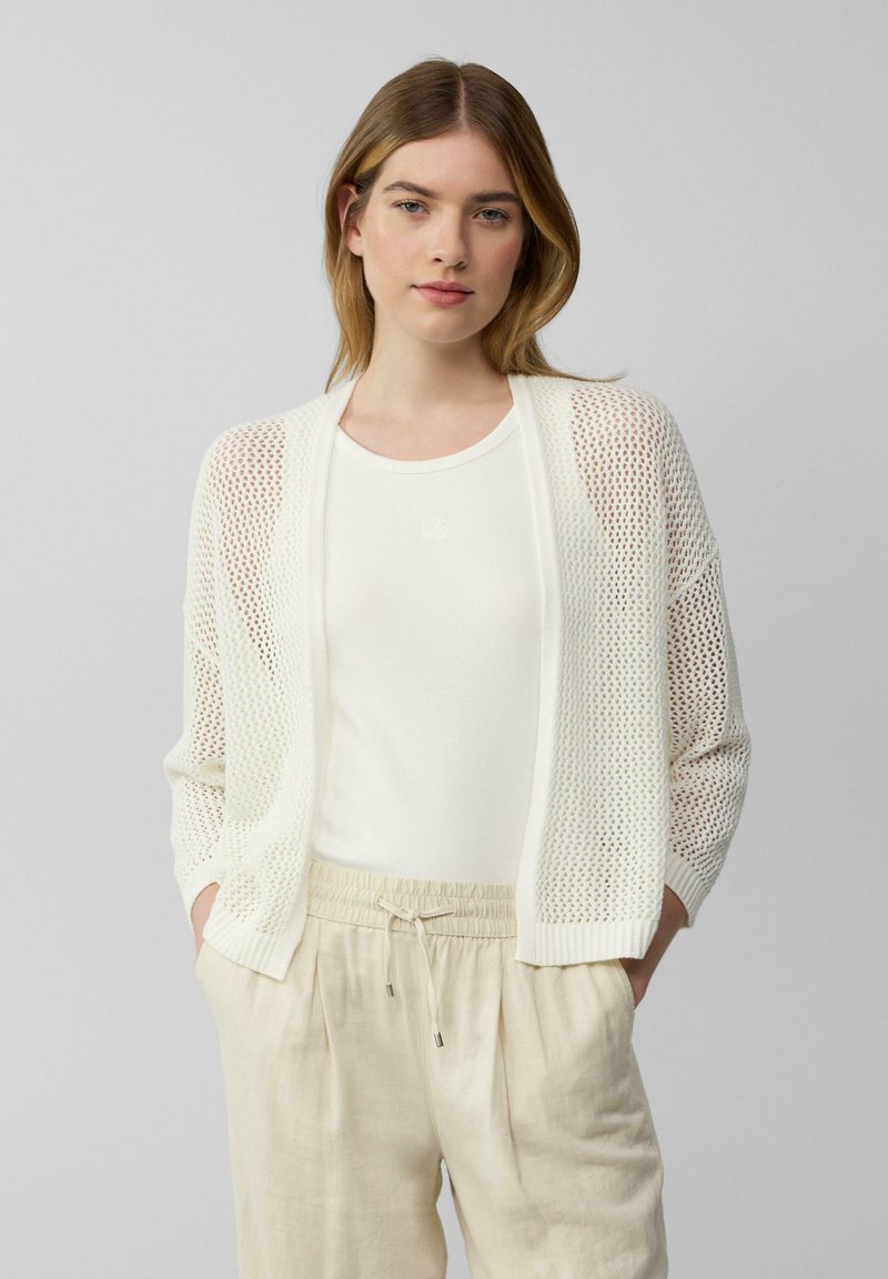 Witte gebreide cardigan met open gaasdesign, gecombineerd met een witte tanktop en lichtbeige broek. Heeft een relaxte pasvorm en ribgebreide manchetten.