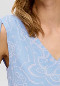 Una donna che indossa un top senza maniche di colore azzurro chiaro con motivi floreali bianchi e ha lunghi capelli castano chiaro mossi che le cadono sulla spalla.