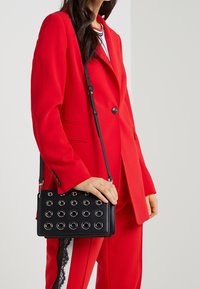 Bolso negro de bandolera con detalles de ojales redondos, forma rectangular, textura suave y correa ajustable, llevado con un traje rojo.