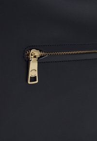 Szczegół czarnej skórzanej torby z złotym zamkiem błyskawicznym i logo "COACH NEW YORK" na suwaku. Gładka tekstura i minimalistyczny design.