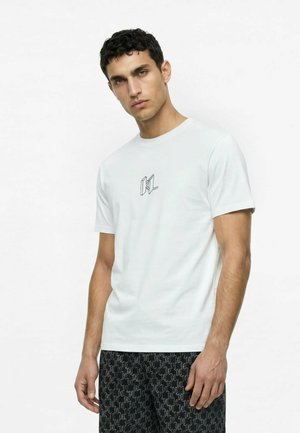 Joven con camiseta blanca de manga corta con pequeño logo negro y pantalones negros con estampado, de pie contra un fondo blanco liso.