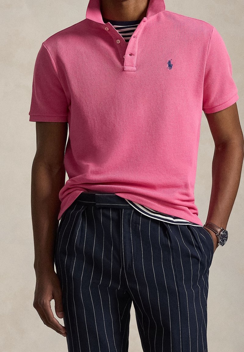 Homme portant un polo rose vif avec un petit logo, un pantalon bleu marine à fines rayures et une montre au poignet gauche.