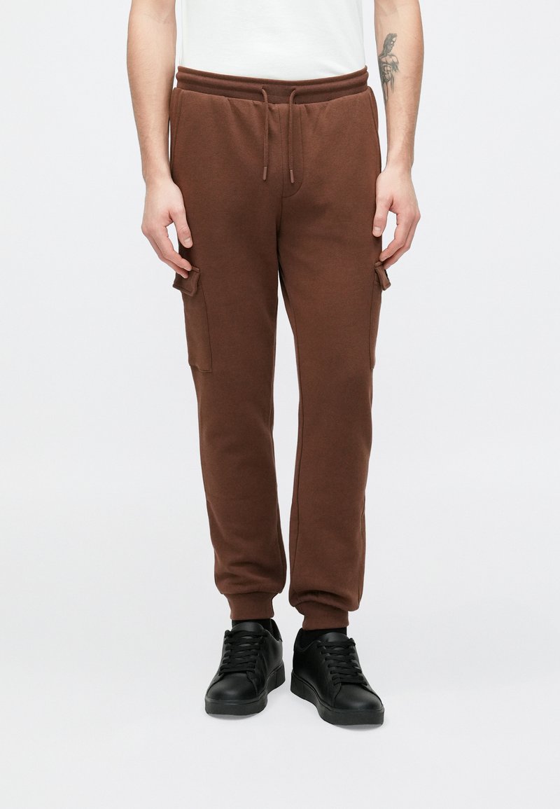 Brune joggingbukser med elastisk talje, snøre og sidelommer med cargo-stil. Bærer sorte sneakers med en glat overflade.