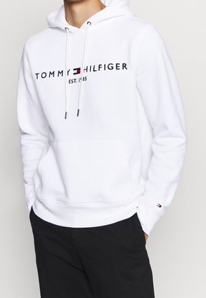 Witte hoodie van zachte stof, met een voorzak in kangoeroestijl, een trekkoordcapuchon en een logo in marineblauw, rood en wit. Zwarte broek zichtbaar.