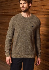 Hnědý texturovaný svetr s kulatým výstřihem a dlouhými rukávy. Má malé černé logo na hrudi. Nosit s tmavými kalhotami.