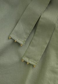 Țesătură de culoare verde măsliniu cu două bretele cusute, fiecare având la capete trei mărgele aurii grupate în clustere mici.