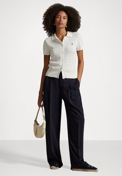 Žena s kudrnatými vlasy má na sobě bílý krátký pletený kardigan s krátkými rukávy, černé široké kalhoty, espadrilky a drží béžovou kabelku na jednoduchém pozadí.