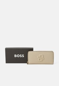Bēšs ādas maks ar reljefotu logotipu, ar rāvējslēdzēju un zelta toni aparatūru. Prezentēts melnā kastē ar baltu tekstu "BOSS."