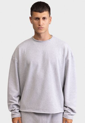 Νεαρός άνδρας με κοντά σκούρα μαλλιά φοράει άνετο ανοιχτό γκρι φούτερ και αντίστοιχο παντελόνι, σε απλό ανοιχτό φόντο.