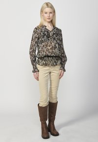 Blusa nera trasparente con stampa floreale, scollo a V e maniche a volant, abbinata a pantaloni in corduroy beige e stivali marroni fino al ginocchio.
