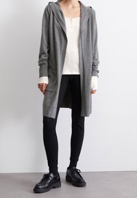 Grå lång cardigan med huva, mjukt tyg, öppen framsida, sidofickor, lager på lager över en vit topp och kombinerad med svarta leggings och skor.