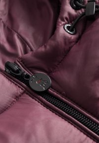 Bordeaux pufferjack stof met glanzende textuur, met een zwarte ritssluiting en een ronde ritshanger met een logo-ontwerp.