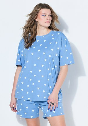 Frau mit welligem Haar, die ein hellblaues Pyjama-Set mit weißen Herzmustern trägt, Ringe an den Fingern und blauen Nagellack hat, schaut nach rechts.