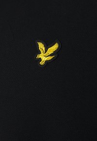 Svart tyg har en gul broderad örnlogo. Texturen är slät med ett lätt vävningsmönster.