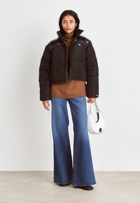 Zwarte pufferjack met glanzende zwarte schouderaccenten, bruine gebreide trui, brede blauwe denim jeans en een witte handtas.