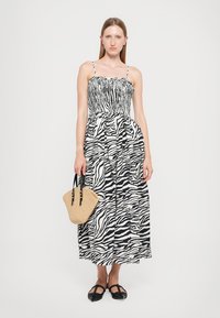 Svart og hvit zebra-striped maxi-kjole med smocket bodice, tynne stropper og sydd skjørt; paret med en vevd beige veske.
