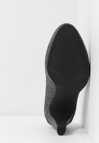 Chaussure à talons pour femme avec semelle noire texturée et dessus à motif gris, reposant contre un mur et un sol blanc.