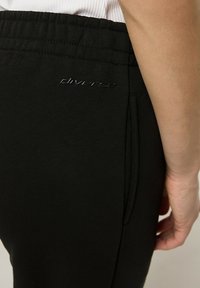 Czarne sportowe spodnie o gładkiej teksturze, elastycznym pasie oraz dyskretnym logo na kieszeni. Posiadają boczną kieszeń na drobne przedmioty.